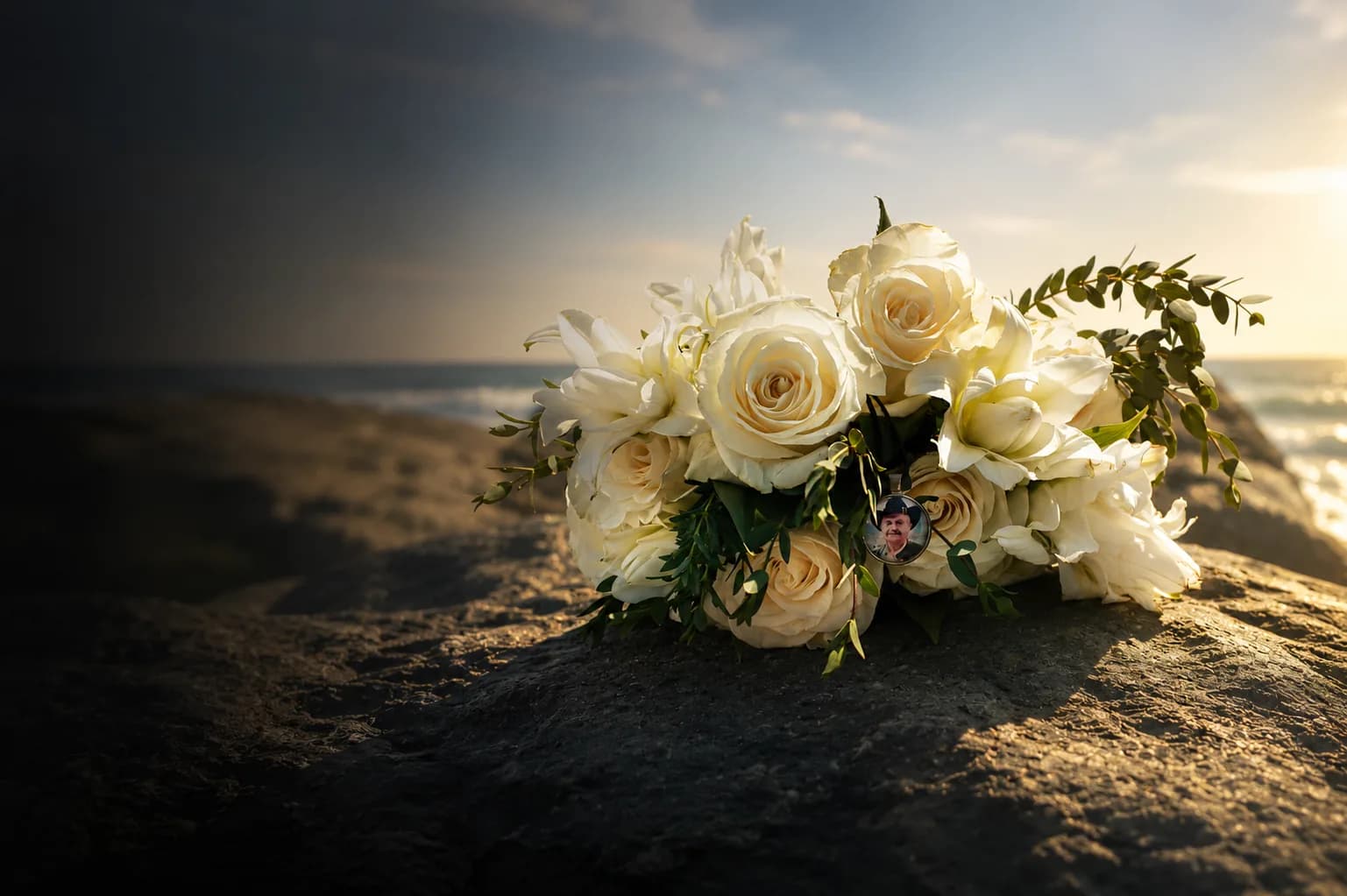 A white rose bouquet with a memorial locket, resting on the sand.