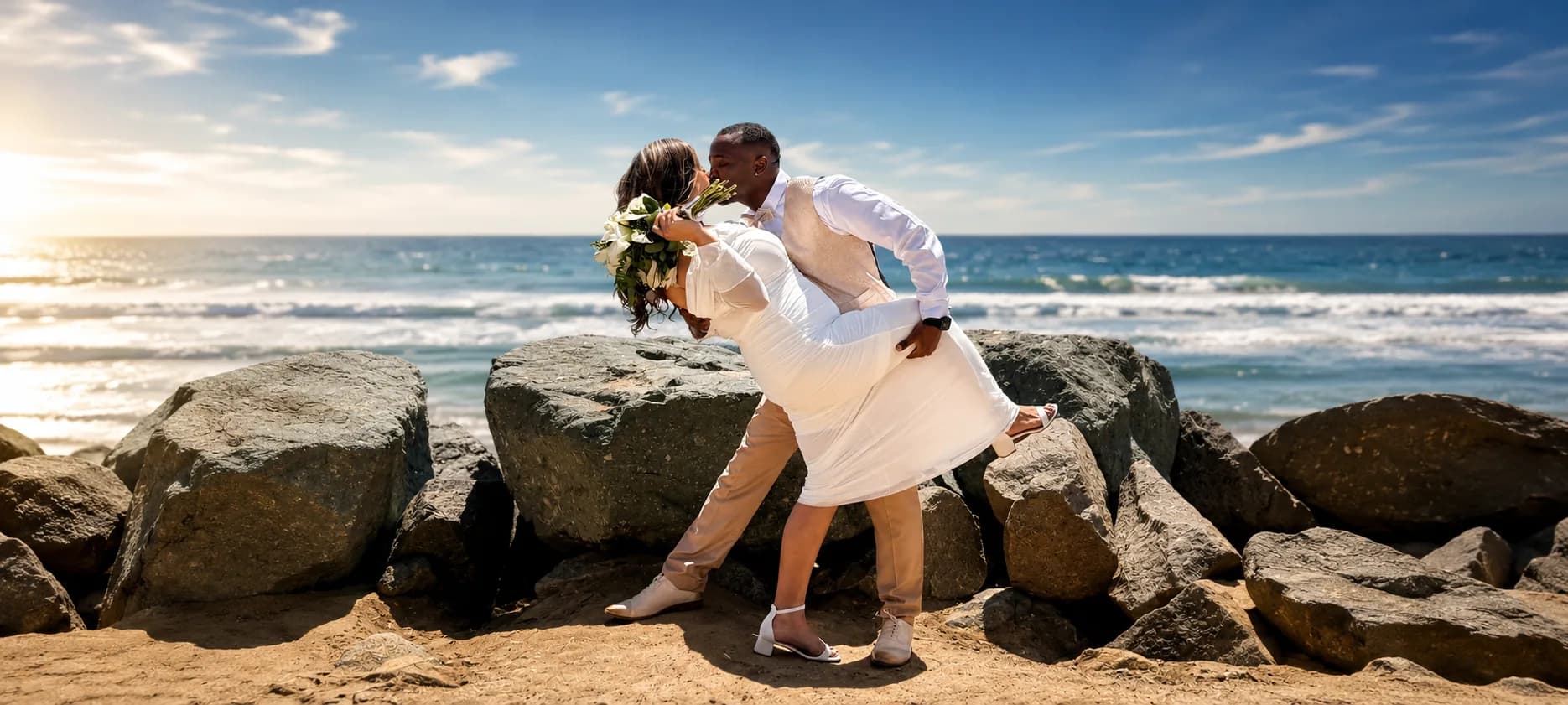 A groom dipping his bride into a kiss on a rocky coastline.